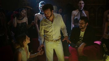 Movie still from “Urge” (2016), directed by Aaron Kaufman – A group of people sitting in a room with a man in a white shirt; Medium shot, Low angle