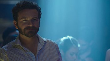 Movie still from “Urge” (2016), directed by Aaron Kaufman – A man standing in a room with people in the background; Close Up shot, Over the shoulder angle