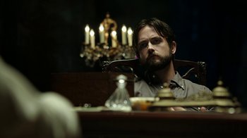 Movie still from “Urge” (2016), directed by Aaron Kaufman – A man sitting at a dinner table with candles in the background; Medium shot, Over the shoulder angle