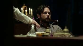 Movie still from “Urge” (2016), directed by Aaron Kaufman – A man sitting in front of a table holding a pipe in his mouth; Medium shot, Over the shoulder angle