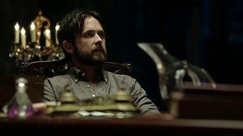 Movie still from “Urge” (2016), directed by Aaron Kaufman – A man sitting at a table with a beard; Close Up shot, Low angle