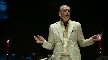 Movie still from “Urge” (2016), directed by Aaron Kaufman – A man in a white suit is standing in front of a black background; Medium shot, Low angle