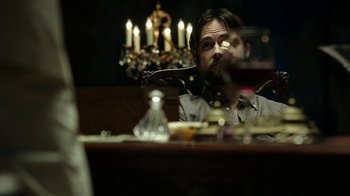 Movie still from “Urge” (2016), directed by Aaron Kaufman – A man sitting at a dinner table looking at the camera; Medium shot, Over the shoulder angle