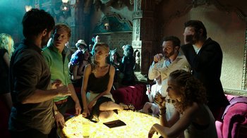 Movie still from “Urge” (2016), directed by Aaron Kaufman – A group of people sitting around a round table; Wide shot, High angle