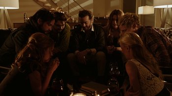 Movie still from “Urge” (2016), directed by Aaron Kaufman – A group of people sitting around a table with wine glasses on it; Medium shot, High angle