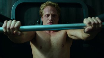 Movie still from “Urge” (2016), directed by Aaron Kaufman – A shirtless man is lifting a barbell in a gym; Close Up shot, Low angle