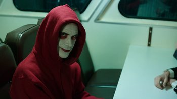 Movie still from “Urge” (2016), directed by Aaron Kaufman – A person in a red hooded sweatshirt sitting on a bench; Medium shot, High angle