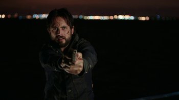Movie still from “Urge” (2016), directed by Aaron Kaufman – A man holding a gun while standing in a field; Close Up shot, Over the shoulder angle