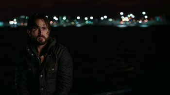 Movie still from “Urge” (2016), directed by Aaron Kaufman – A man standing in front of a body of water at night; Close Up shot, Over the shoulder angle