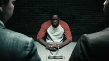 Movie still from “Snowfall” (2017), directed by Hiro Murai – A man chained to a table in a jail cell; Medium shot, High angle