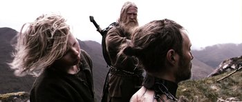 Movie still from “Valhalla Rising” (2009), directed by Nicolas Winding Refn – A man with long hair and a beard is standing next to two other men; Medium shot, Over the shoulder angle