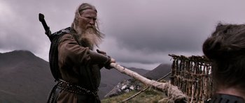 Movie still from “Valhalla Rising” (2009), directed by Nicolas Winding Refn – A man with a long beard is holding a wooden stick; Medium shot, Low angle
