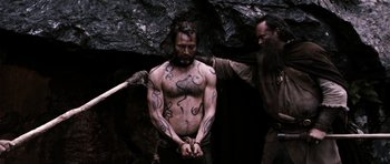 Movie still from “Valhalla Rising” (2009), directed by Nicolas Winding Refn – A man with a beard is tied to a rope; Medium shot, Over the shoulder angle