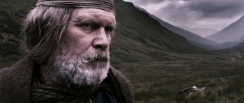 Movie still from “Valhalla Rising” (2009), directed by Nicolas Winding Refn – An old man with a beard and a long white beard is standing in front of a mountain; Close Up shot, Low angle