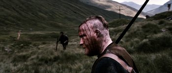 Movie still from “Valhalla Rising” (2009), directed by Nicolas Winding Refn – A man with a face tattoo is walking in a field; Close Up shot, Over the shoulder angle