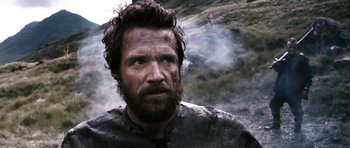 Movie still from “Valhalla Rising” (2009), directed by Nicolas Winding Refn – A man with a long beard is standing in front of a mountain; Close Up shot, Low angle