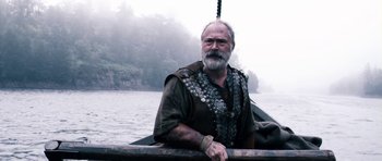 Movie still from “Valhalla Rising” (2009), directed by Nicolas Winding Refn – An older man with a beard is sitting on a boat in the middle of a river; Medium shot, Low angle
