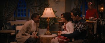 Movie still from “Violent Night” (2022), directed by Tommy Wirkola – A group of people sitting around a table with a lamp on it; Medium shot, Over the shoulder angle