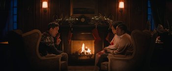 Movie still from “Violent Night” (2022), directed by Tommy Wirkola – A group of people sitting in front of a fireplace; Medium shot, Over the shoulder angle