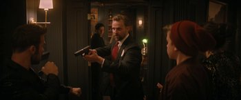 Movie still from “Violent Night” (2022), directed by Tommy Wirkola – A man in a suit and tie holding a gun in his hand; Medium shot, Over the shoulder angle