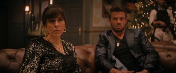 Movie still from “Violent Night” (2022), directed by Tommy Wirkola – A woman and a man sitting on a couch in a room; Medium shot, Over the shoulder angle