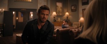 Movie still from “Violent Night” (2022), directed by Tommy Wirkola – A man sitting in front of another man in a living room; Medium shot, Over the shoulder angle