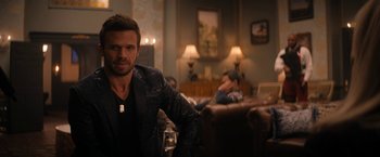 Movie still from “Violent Night” (2022), directed by Tommy Wirkola – A man sitting in front of two other men in a living room; Close Up shot, Over the shoulder angle