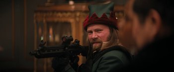 Movie still from “Violent Night” (2022), directed by Tommy Wirkola – A man with a beard wearing a red and green hat and holding a rifle; Close Up shot, High angle