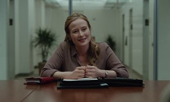 Movie still from “Vox Lux” (2018), directed by Brady Corbet – A woman sitting at a table in an office setting; Medium shot, Over the shoulder angle