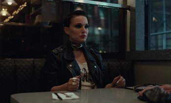 Movie still from “Vox Lux” (2018), directed by Brady Corbet – A woman sitting at a table in front of cigarettes; Medium shot, Over the shoulder angle