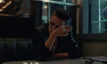 Movie still from “Vox Lux” (2018), directed by Brady Corbet – A person sitting at a table with a cell phone in their hand; Medium shot, Over the shoulder angle