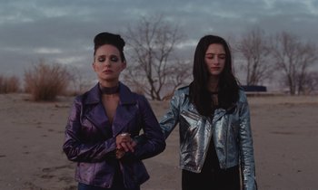 Movie still from “Vox Lux” (2018), directed by Brady Corbet – Two young women holding hands while standing on a dirt road; Medium shot, Low angle