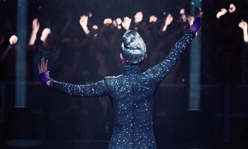 Movie still from “Vox Lux” (2018), directed by Brady Corbet – A person in a black outfit raising their hands in front of a crowd of people; Medium shot, Over the shoulder angle