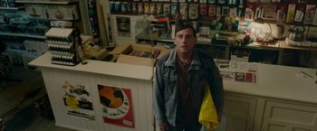 Movie still from “Welcome to Marwen” (2018), directed by Robert Zemeckis – A man standing in front of a store holding a yellow bag; Medium shot, High angle