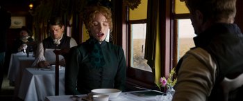 Movie still from “Woman Walks Ahead” (2017), directed by Susanna White – A woman sitting at a table with a plate in front of a window; Medium shot, Low angle