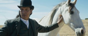 Movie still from “Woman Walks Ahead” (2017), directed by Susanna White – A man wearing a top hat is petting a white horse; Close Up shot, Over the shoulder angle