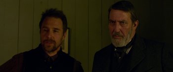 Movie still from “Woman Walks Ahead” (2017), directed by Susanna White – Two men standing next to each other in front of a wall; Close Up shot, Over the shoulder angle