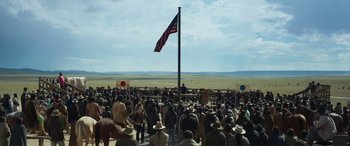 Movie still from “Woman Walks Ahead” (2017), directed by Susanna White – A crowd of people standing under a large american flag; Extreme Wide shot, Over the shoulder angle