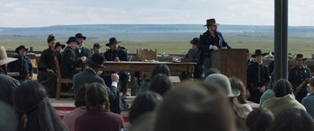 Movie still from “Woman Walks Ahead” (2017), directed by Susanna White – A man is speaking at an event with a group of people watching; Wide shot, Over the shoulder angle