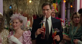 Movie still from “Working Girl” (1988), directed by Mike Nichols – A man in a suit and tie drinking a drink; Medium shot, Over the shoulder angle
