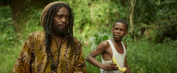 Movie still from “Yardie” (2018), directed by Idris Elba – A man and a boy in the grass with bananas; Medium shot, Over the shoulder angle