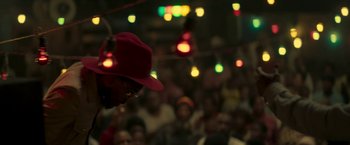 Movie still from “Yardie” (2018), directed by Idris Elba – A crowd of people sitting in a room with lights in the background; Close Up shot, Over the shoulder angle