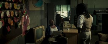 Movie still from “Yardie” (2018), directed by Idris Elba – A man sitting in a chair in front of a television; Medium shot, High angle