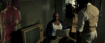 Movie still from “Yardie” (2018), directed by Idris Elba – A man sitting in front of a television holding a newspaper; Medium shot, High angle