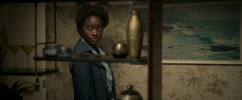 Movie still from “Yardie” (2018), directed by Idris Elba – A woman standing in front of a shelf in a room; Medium shot, Over the shoulder angle