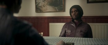 Movie still from “Yardie” (2018), directed by Idris Elba – A man sitting at a table in front of a painting; Medium shot, Over the shoulder angle