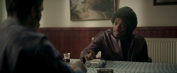 Movie still from “Yardie” (2018), directed by Idris Elba – A man sitting at a table with a remote control in his hand; Close Up shot, Over the shoulder angle