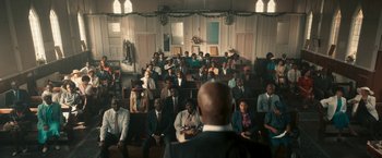 Movie still from “Yardie” (2018), directed by Idris Elba – A group of people sitting in a room with a man standing in front of them; Extreme Wide shot, High angle