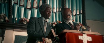 Movie still from “Yardie” (2018), directed by Idris Elba – Two men in suits and ties sitting in front of a book; Medium shot, Low angle