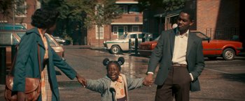 Movie still from “Yardie” (2018), directed by Idris Elba – A little girl with mickey mouse ears is holding hands with a man and woman; Medium shot, High angle